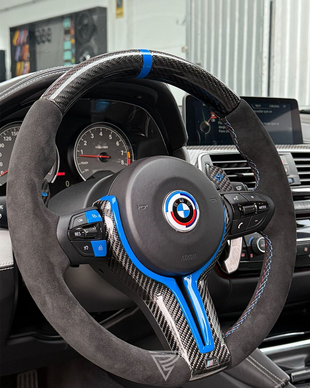 Volante de un BMW M forrado en alcántara, con el aro y los radios en fibra de carbono, insertos azules y costura tricolor, con el cuadro de relojes detrás