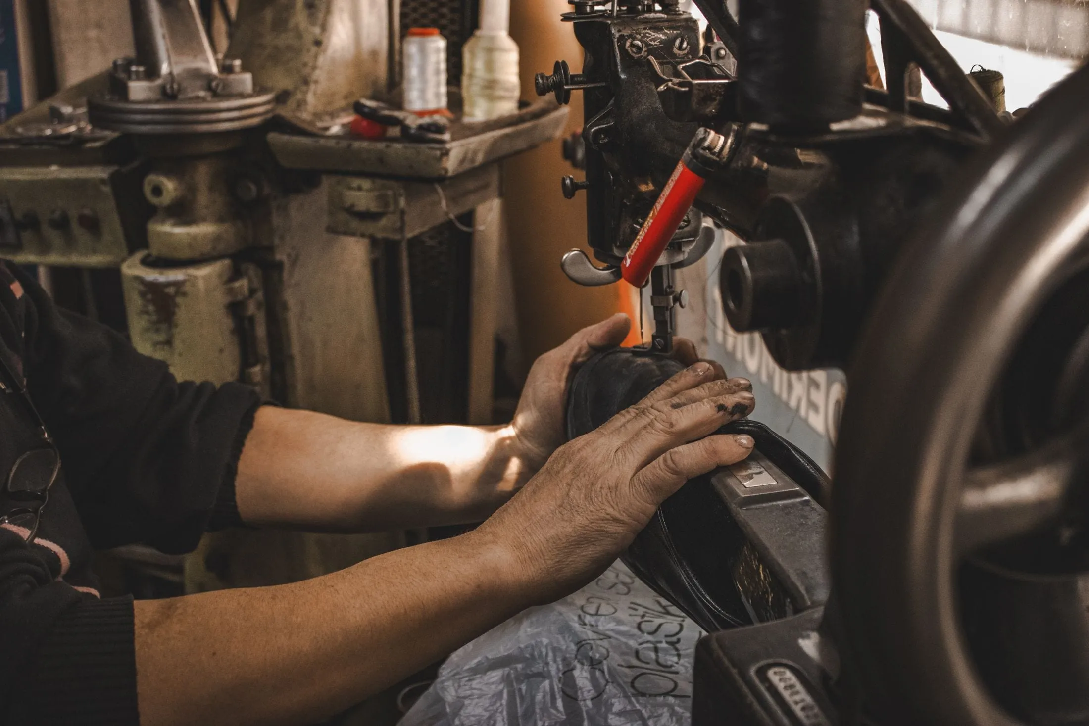 Manos guiando una pieza de piel oscura bajo el prensatelas de una máquina de coser de brazo, en un taller con carretes de hilo al fondo