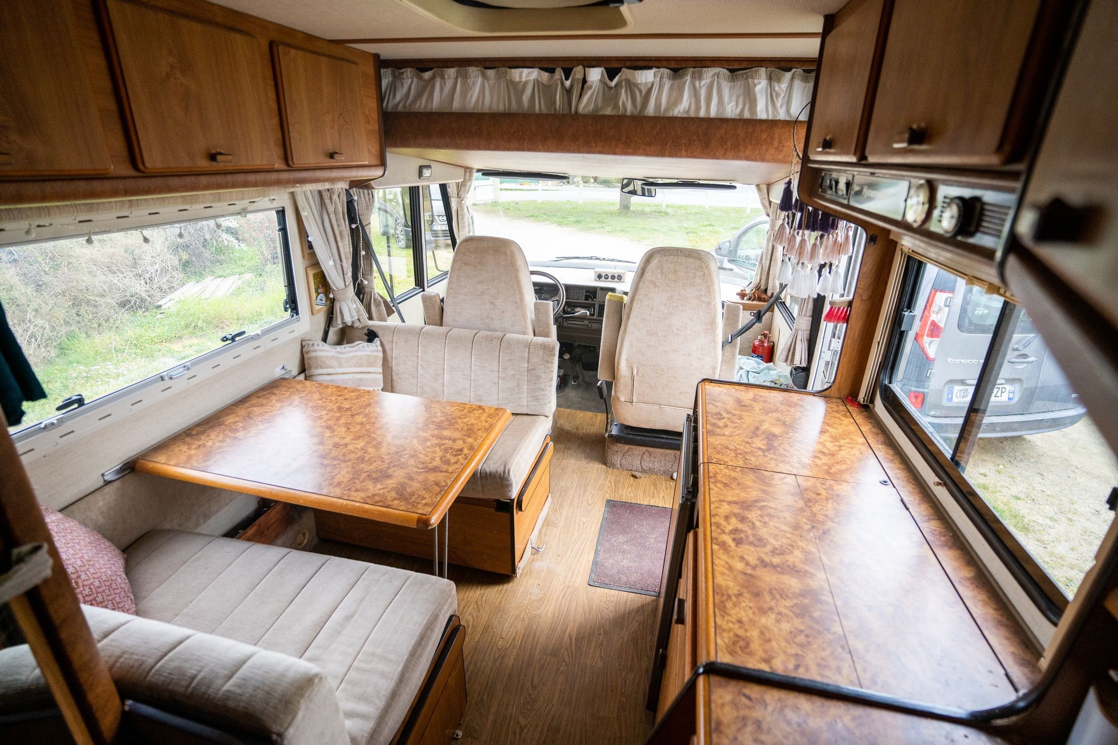 Interior de una camper con el banco y la mesa de madera vistos desde la puerta lateral, con los cojines tapizados en tela clara