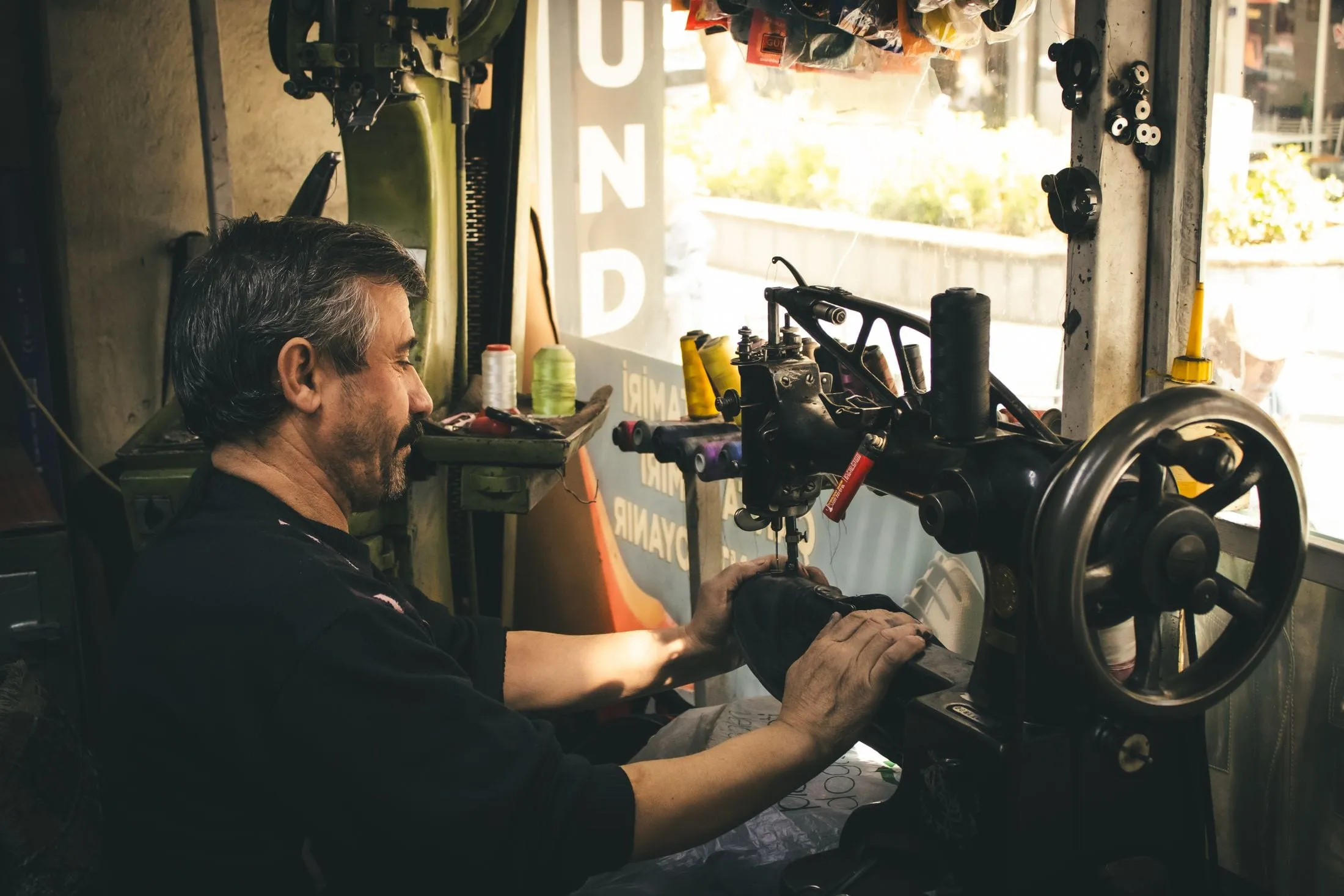 Tapicero cosiendo una pieza de piel en una máquina de coser de brazo, junto a la ventana de su taller
