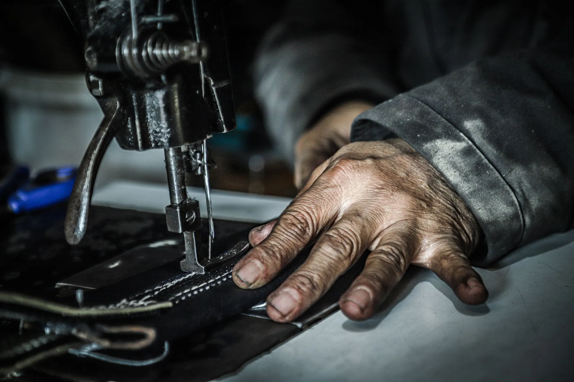 Primer plano de una mano guiando una pieza de piel negra bajo el prensatelas de una máquina de coser industrial, con la costura ya rematada